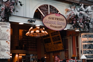 A charming storefront of a sandwich shop, adorned with an array of flowers and greenery above the entrance. The sign reads 'Gaufreirie Maison Hector Sandwicherie,' with another sign detailing menu items like 'gaufres' and 'beignets.' The warm, inviting atmosphere features a vintage-style chandelier and various menu boards in French.