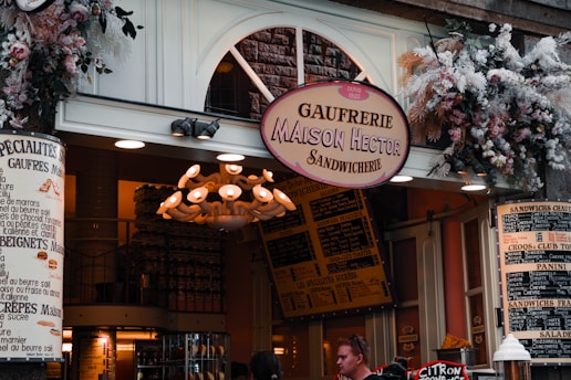 A charming storefront of a sandwich shop, adorned with an array of flowers and greenery above the entrance. The sign reads 'Gaufreirie Maison Hector Sandwicherie,' with another sign detailing menu items like 'gaufres' and 'beignets.' The warm, inviting atmosphere features a vintage-style chandelier and various menu boards in French.