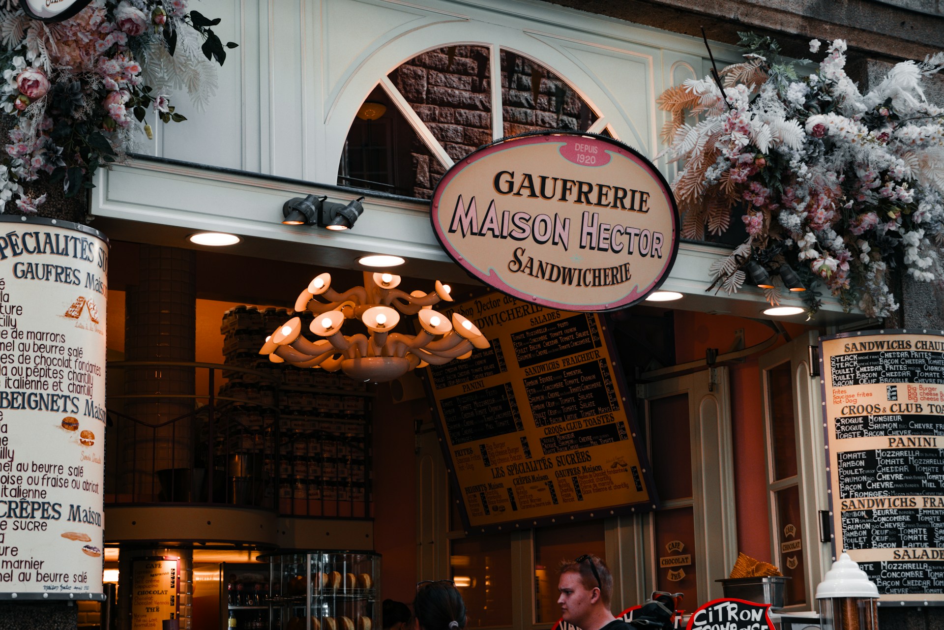 A charming storefront of a sandwich shop, adorned with an array of flowers and greenery above the entrance. The sign reads 'Gaufreirie Maison Hector Sandwicherie,' with another sign detailing menu items like 'gaufres' and 'beignets.' The warm, inviting atmosphere features a vintage-style chandelier and various menu boards in French.