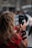 A person with long, wavy hair wearing a red shirt is holding a smartphone with both hands, taking a picture or video. The phone is partially covered by a case displaying travel-related stickers. The background appears blurred, suggesting a busy outdoor setting with several people.