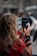 A person with long, wavy hair wearing a red shirt is holding a smartphone with both hands, taking a picture or video. The phone is partially covered by a case displaying travel-related stickers. The background appears blurred, suggesting a busy outdoor setting with several people.