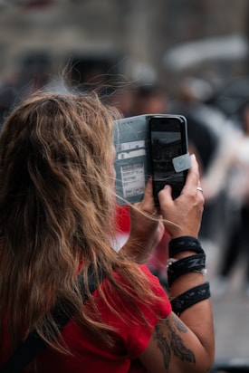 A person with long, wavy hair wearing a red shirt is holding a smartphone with both hands, taking a picture or video. The phone is partially covered by a case displaying travel-related stickers. The background appears blurred, suggesting a busy outdoor setting with several people.