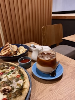 A wooden table is set with a spread of food, including a pizza topped with various vegetables like olives, cherry tomatoes, and mushrooms. Next to it is a blue saucer holding a clear glass of iced coffee with a layer of milk. There is also a blue bowl containing a grilled sandwich and potato chips, accompanied by a small cup of tomato sauce and a white creamer.