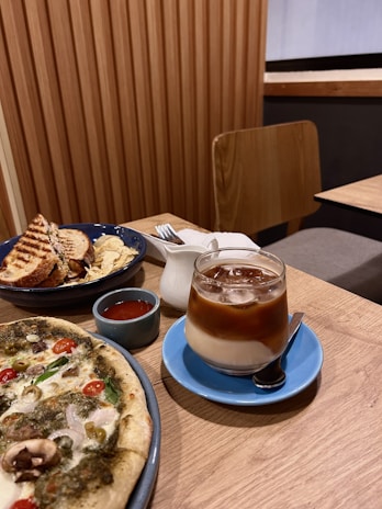 A wooden table is set with a spread of food, including a pizza topped with various vegetables like olives, cherry tomatoes, and mushrooms. Next to it is a blue saucer holding a clear glass of iced coffee with a layer of milk. There is also a blue bowl containing a grilled sandwich and potato chips, accompanied by a small cup of tomato sauce and a white creamer.