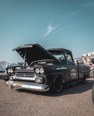 A vintage black Chevrolet truck with its hood open, parked outdoors on an asphalt surface. The words 'Peggy's Chop Shop' are painted on the side of the vehicle. There are other classic cars visible in the background, along with a sky featuring scattered clouds.