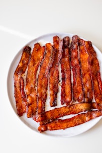 Crispy strips of seasoned bacon are neatly arranged on a clean white plate. The bacon is cooked to a deep brown hue with visible specks of seasoning, suggesting a flavorful preparation.