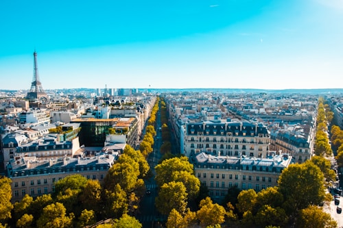 An aerial view of a cityscape featuring a prominent structure resembling the Eiffel Tower on the left. The scene captures numerous classic French-style buildings lined along streets with vibrant green trees. The sky is clear and blue, adding depth to the skyline.