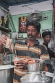 A man wearing a striped shirt is pouring a beverage, likely tea, from a metal container into a glass. The scene appears to be set in a small, modest tea shop or café. Another person is standing in the background, smiling. The surroundings include various items such as jars, a fan, and a wall poster.