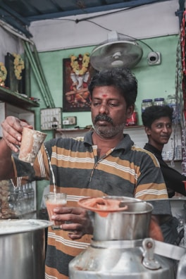 A man wearing a striped shirt is pouring a beverage, likely tea, from a metal container into a glass. The scene appears to be set in a small, modest tea shop or café. Another person is standing in the background, smiling. The surroundings include various items such as jars, a fan, and a wall poster.