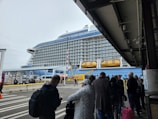 A large cruise ship docked at a terminal with people standing in line and waiting. The ship has multiple decks and lifeboats visible. The weather appears overcast, and the terminal area is partially covered. The people in line are wearing various types of outerwear.