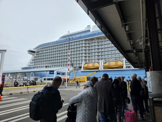 A large cruise ship docked at a terminal with people standing in line and waiting. The ship has multiple decks and lifeboats visible. The weather appears overcast, and the terminal area is partially covered. The people in line are wearing various types of outerwear.
