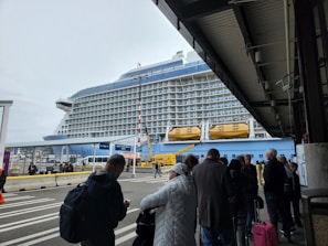 A large cruise ship docked at a terminal with people standing in line and waiting. The ship has multiple decks and lifeboats visible. The weather appears overcast, and the terminal area is partially covered. The people in line are wearing various types of outerwear.