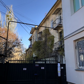 A residential street scene with buildings featuring balconies covered in greenery. A black metal gate with decorative patterns is in the foreground, while multiple electrical wires run overhead. The sky is clear and blue, adding a serene backdrop to the urban environment.