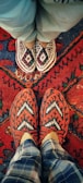 Two pairs of feet wearing intricately patterned, handcrafted slippers are positioned on a vibrant, red patterned rug. The top pair of slippers features a red, white, and blue diamond design, while the bottom pair is red and white with a chevron pattern. The background shows a traditional rug with various geometric designs and bold colors.