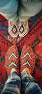 Two pairs of feet wearing intricately patterned, handcrafted slippers are positioned on a vibrant, red patterned rug. The top pair of slippers features a red, white, and blue diamond design, while the bottom pair is red and white with a chevron pattern. The background shows a traditional rug with various geometric designs and bold colors.