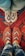 Two pairs of feet wearing intricately patterned, handcrafted slippers are positioned on a vibrant, red patterned rug. The top pair of slippers features a red, white, and blue diamond design, while the bottom pair is red and white with a chevron pattern. The background shows a traditional rug with various geometric designs and bold colors.