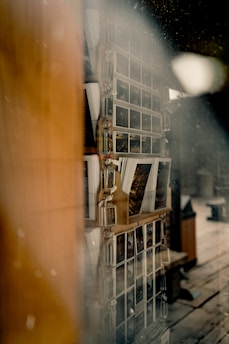 A display rack with multiple rows of postcards is visible through a slightly fogged or dusty glass. The postcards are arranged neatly, with each row containing various images, possibly depicting scenic or artistic photos. There is a warm, wooden frame around the display and the surrounding area hints at a rustic interior setting.