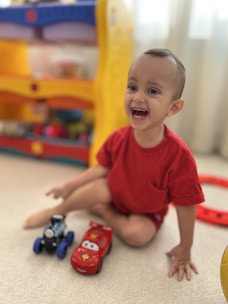 A young child is sitting on a light-colored carpet, smiling joyfully while wearing a red shirt. In front of the child are two toy cars, one red with a character face and the other black with wheels. In the background, there is a colorful toy shelf with yellow and blue elements, partially blurred.
