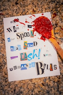 A piece of paper on a granite surface with a collage-style ransom note composed of various cut-out letters. The note mentions sending cash in small, unmarked bills. Red sauce is smeared on the paper, with a wooden spoon covered in sauce resting on top.