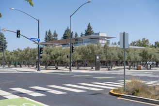 A modern building with a glass facade, surrounded by neatly arranged trees and visible traffic lights at an intersection. The structure includes a prominent logo of a well-known tech company. The street sign reads 'Pruneridge Ave'. There are crosswalks and a bike lane marked on the road, indicating an urban environment with a focus on pedestrian and cyclist traffic.