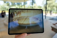 A hand is holding a tablet displaying an augmented reality scene. The tablet screen shows a virtual, circular architectural structure with people standing around it in an outdoor park setting. The background includes blurred figures and trees visible through large windows.