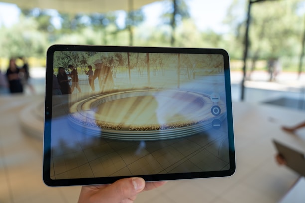 A hand is holding a tablet displaying an augmented reality scene. The tablet screen shows a virtual, circular architectural structure with people standing around it in an outdoor park setting. The background includes blurred figures and trees visible through large windows.