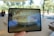 A hand is holding a tablet displaying an augmented reality scene. The tablet screen shows a virtual, circular architectural structure with people standing around it in an outdoor park setting. The background includes blurred figures and trees visible through large windows.