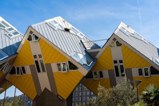 A row of uniquely designed cube-shaped houses with slanted roofs, characterized by their yellow and gray panels. The architectural style is modern and distinctive, giving an impression of innovation and creativity. The sky is clear with a few wispy clouds, and there is some greenery in the foreground.