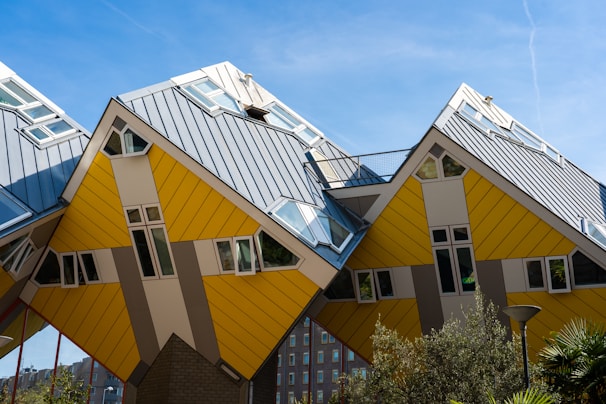 A row of uniquely designed cube-shaped houses with slanted roofs, characterized by their yellow and gray panels. The architectural style is modern and distinctive, giving an impression of innovation and creativity. The sky is clear with a few wispy clouds, and there is some greenery in the foreground.