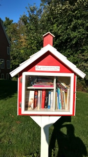 A small, red wooden box resembling a miniature house stands on a white post, serving as a free library. It is filled with various books visible through a glass window. The background includes green grass and dense trees, alongside part of a red brick building.