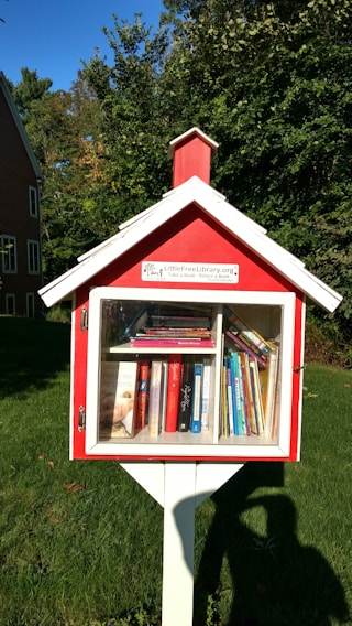 A small, red wooden box resembling a miniature house stands on a white post, serving as a free library. It is filled with various books visible through a glass window. The background includes green grass and dense trees, alongside part of a red brick building.