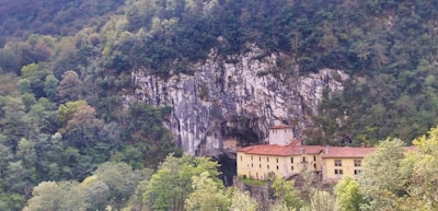 A large monastery or building is nestled into the side of a rocky cliff surrounded by lush green trees. The structure has a red-tiled roof and beige walls, blending with the natural colors of the landscape. The cliff face is rugged and the surrounding forest is dense, creating a sense of seclusion and tranquility.
