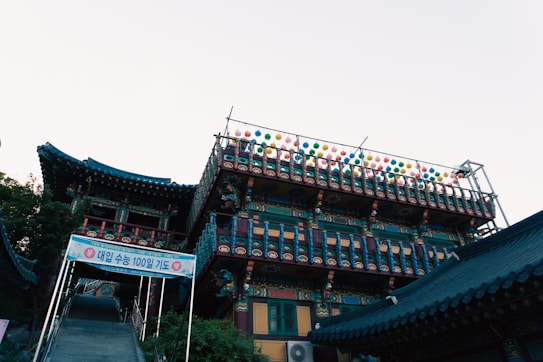 A traditional East Asian building with ornate architectural details, featuring a multilevel structure adorned with colorful lanterns hanging from the upper balcony. The roof is decorated with intricate patterns and vibrant colors. A banner with Korean text hangs above a staircase leading to the entrance.