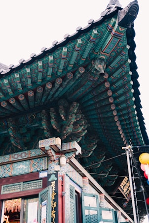 A close-up view of an intricately decorated temple roof with detailed wooden carvings and vibrant traditional patterns. The building features colorful paintwork in shades of teal, red, and gold, complemented by ornate brackets and wooden beams. A hanging lantern and signage with Asian script are visible near the entrance.