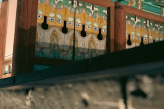 An intricately designed wooden structure features artistic and colorful patterns with motifs resembling faces or masks, incorporating blues, greens, and golds. The design reflects traditional or cultural influence and is framed by wooden beams.