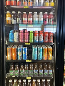 A refrigerated display case holds several rows of bottled and canned beverages. The top shelf contains a variety of flavored Vitaminwater in different colors, such as orange, red, pink, and clear. The middle shelves feature rows of drinks like Prime Hydration and colorful Peace Tea cans with artistic designs. The bottom shelves display bottles of Pure Leaf iced tea in brown, clear plastic containers. Each shelf has small price labels indicating the cost of the drinks.