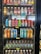 A refrigerated display case holds several rows of bottled and canned beverages. The top shelf contains a variety of flavored Vitaminwater in different colors, such as orange, red, pink, and clear. The middle shelves feature rows of drinks like Prime Hydration and colorful Peace Tea cans with artistic designs. The bottom shelves display bottles of Pure Leaf iced tea in brown, clear plastic containers. Each shelf has small price labels indicating the cost of the drinks.
