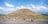 A large stone pyramid with a stepped design dominates the center of a wide open area. Several groups of people are scattered around the base of the pyramid, some appearing to be tourists exploring the site. The sky above is mostly clear with a few fluffy clouds, suggesting a pleasant day. Surroundings include grassy areas and distant hills.