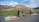 Four people wearing hats are selling colorful handicrafts and textiles on the side of a road, set against the backdrop of an ancient pyramid and lush green landscape.
