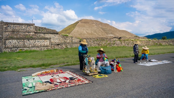 Four people wearing hats are selling colorful handicrafts and textiles on the side of a road, set against the backdrop of an ancient pyramid and lush green landscape.