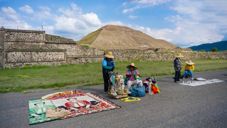 Four people wearing hats are selling colorful handicrafts and textiles on the side of a road, set against the backdrop of an ancient pyramid and lush green landscape.