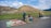 Four people wearing hats are selling colorful handicrafts and textiles on the side of a road, set against the backdrop of an ancient pyramid and lush green landscape.