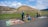 Four people wearing hats are selling colorful handicrafts and textiles on the side of a road, set against the backdrop of an ancient pyramid and lush green landscape.