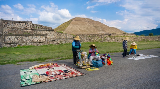 Four people wearing hats are selling colorful handicrafts and textiles on the side of a road, set against the backdrop of an ancient pyramid and lush green landscape.