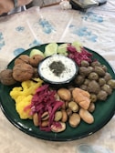 A green plate is filled with a variety of foods including pickles, large beans, olives, cauliflower, and walnuts. In the center, there's a small bowl of yogurt topped with herbs. The arrangement is colorful with a rustic presentation.