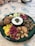 A green plate is filled with a variety of foods including pickles, large beans, olives, cauliflower, and walnuts. In the center, there's a small bowl of yogurt topped with herbs. The arrangement is colorful with a rustic presentation.