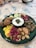 A green plate is filled with a variety of foods including pickles, large beans, olives, cauliflower, and walnuts. In the center, there's a small bowl of yogurt topped with herbs. The arrangement is colorful with a rustic presentation.