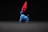 A brightly colored, handcrafted figurine with exaggerated features. The body is predominantly blue with circular patterns, and it has large, vibrant red ears. The figure sits against a dark background, creating a striking contrast that highlights its vivid colors and unique design.