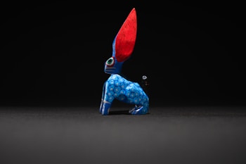 A brightly colored, handcrafted figurine with exaggerated features. The body is predominantly blue with circular patterns, and it has large, vibrant red ears. The figure sits against a dark background, creating a striking contrast that highlights its vivid colors and unique design.