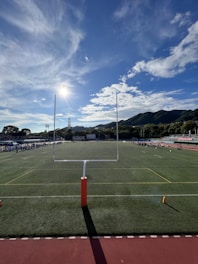 A sports field with green grass and a goalpost is in the foreground. In the distance, a group of people is gathered by the sideline. There are mountains in the background under a blue sky with scattered clouds. The sun is shining brightly, casting long shadows across the field.